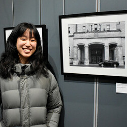 Annie, a young artist, smiles while standing next to her framed black-and-white photograph displayed at an art exhibition. The photograph showcases an architectural building with arched entrances and a parked car in the foreground.