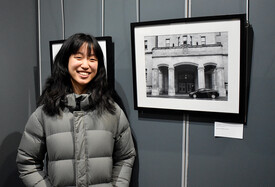 Annie, a young artist, smiles while standing next to her framed black-and-white photograph displayed at an art exhibition. The photograph showcases an architectural building with arched entrances and a parked car in the foreground.