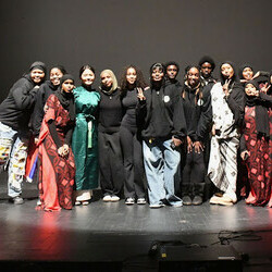 A diverse group of performers and organizers stand together on a stage, dressed in a mix of modern and traditional attire, celebrating Black History Month. Some are wearing flowing red and black patterned garments, while others are in casual and stylish o