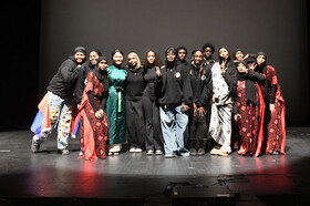 A diverse group of performers and organizers stand together on a stage, dressed in a mix of modern and traditional attire, celebrating Black History Month. Some are wearing flowing red and black patterned garments, while others are in casual and stylish o